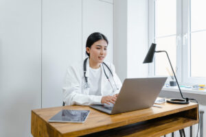 A doctor in laboratory coat typing on a laptop behind a wooden desk